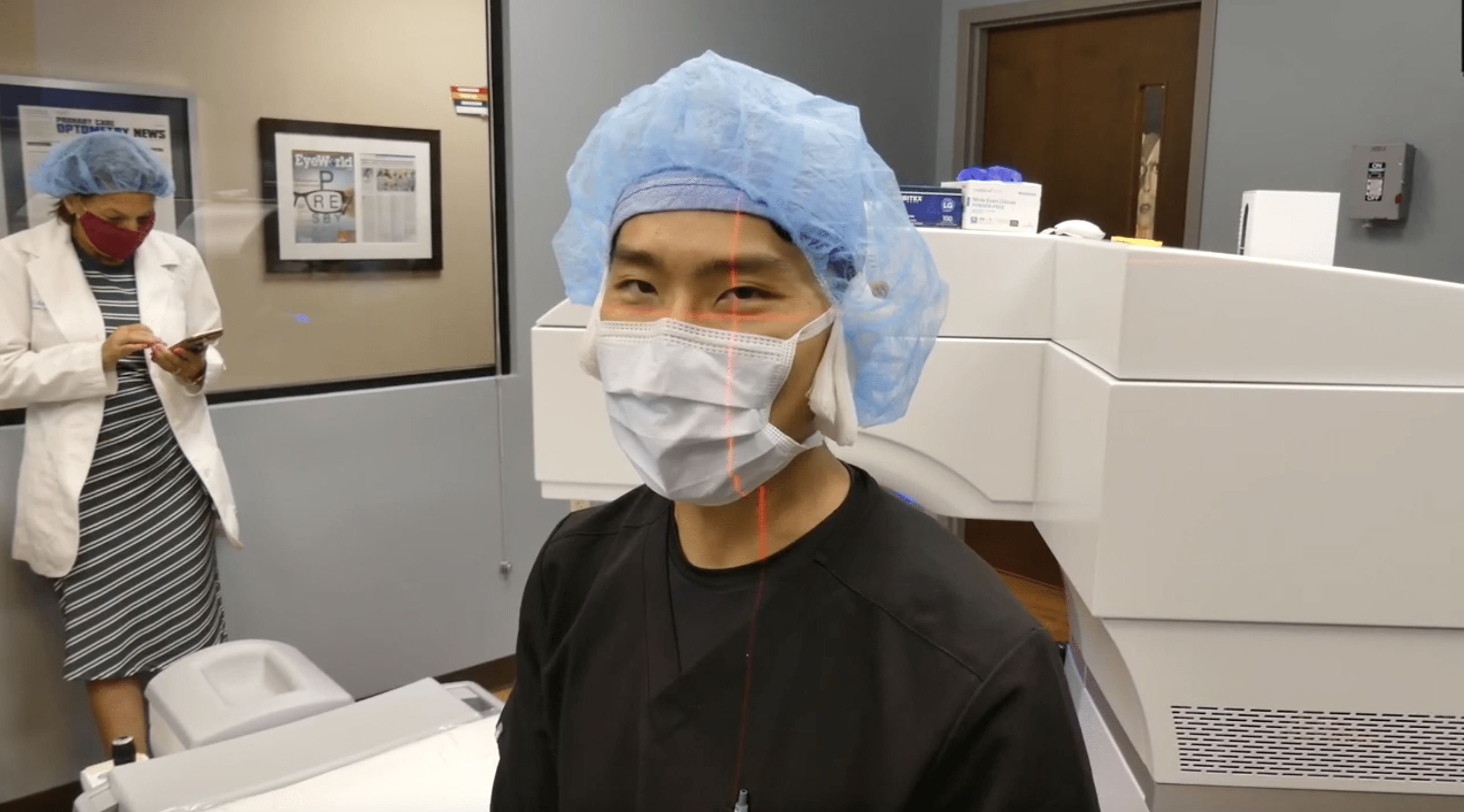 Dr. Joseph Ling wearing a blue surgical cap, face mask, and black scrubs sitting in front of an eye surgery machine