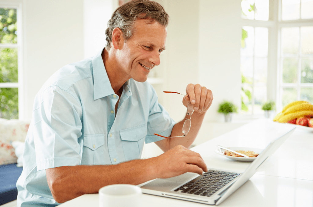 A middle-aged man smiling while sitting in front of his laptop, engaged in a virtual Custom Lens Replacement (CLR) consultation